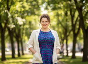 Pewna siebie kobieta plus size w kolorowej sukience i lekkiej marynarce stoi w parku miejskim.